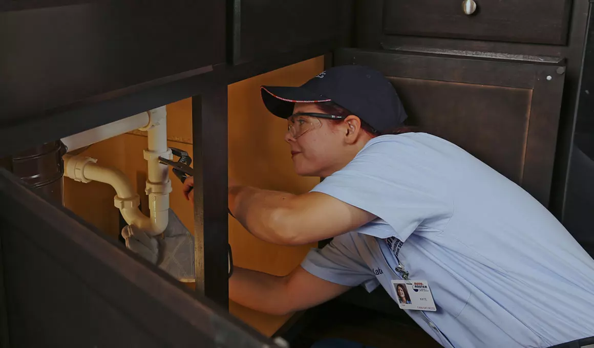 Female plumber wearing a navy baseball cap and light blue shirt using a wrench on a pipe under sink.