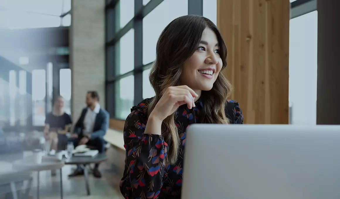 Woman with long wavy hair sitting in front of laptop in a lobby, looking away from the camera, smiling. She is in focus while the people in the background are out of focus.