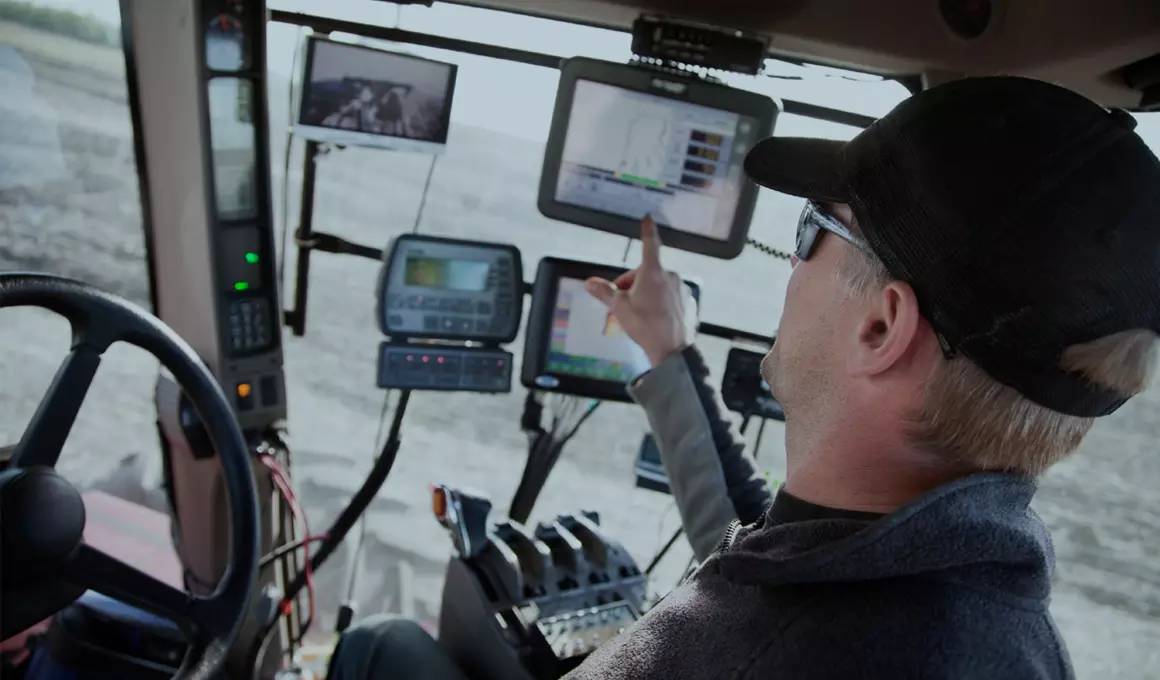 Middle-aged man sitting inside a large piece of farming equipment. He is facing away, pointing to one for 2 touch screens.